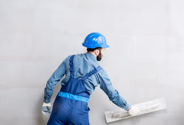 Bauarbeiter mit blauem Helm und blauer Arbeitskleidung, der eine Glättkelle an einer Wand verwendet.