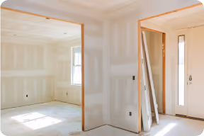 Unfertiger Innenraum eines Hauses mit weißen Trockenbauwänden, Fenstern und einer Tür, Sonnenlicht fällt durch die Fenster.