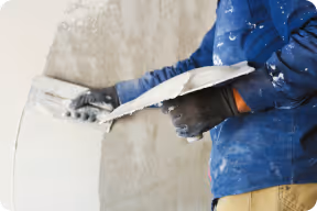 Handwerker trägt blaue Kleidung und glättet Putz mit Kelle an einer Wand.